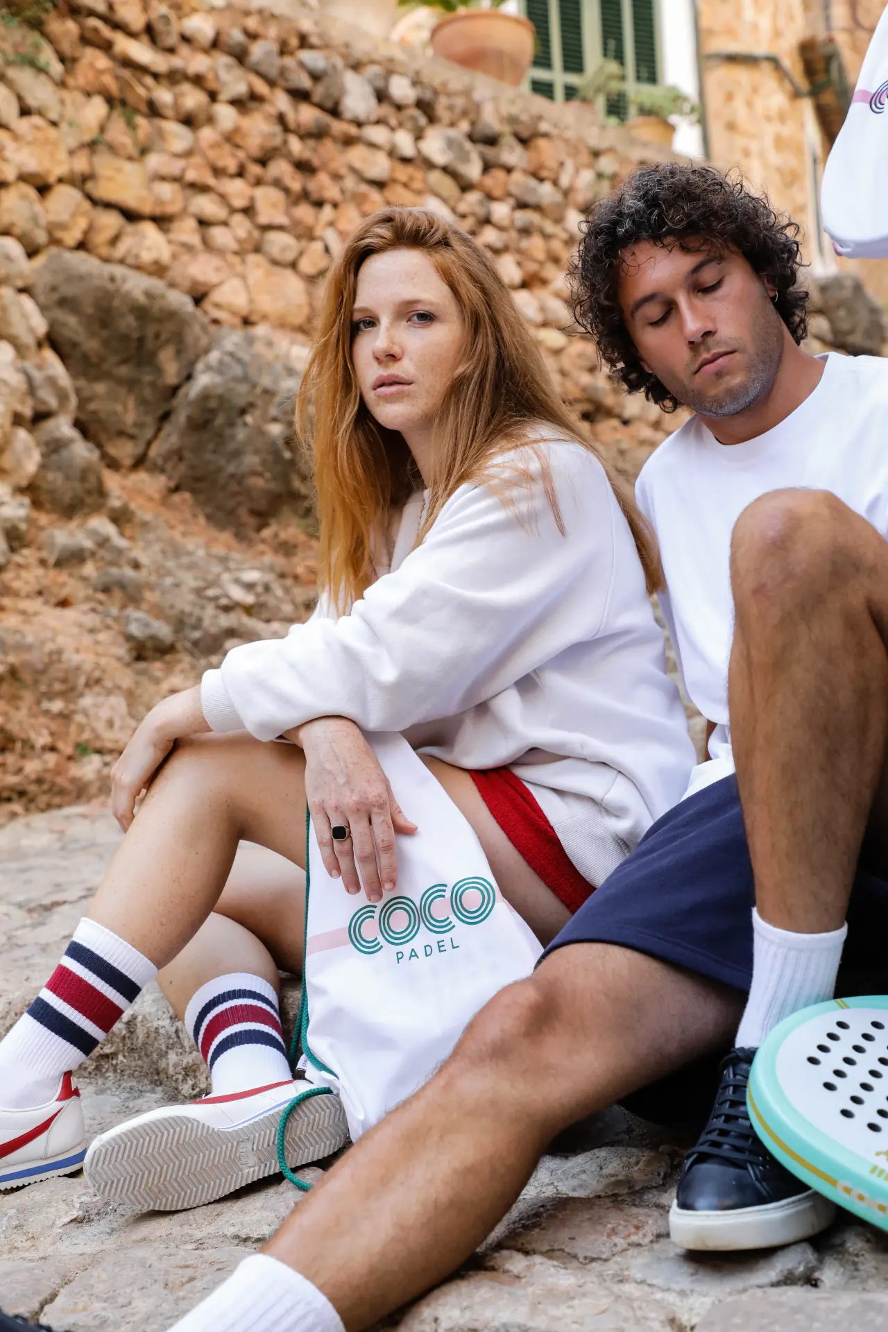 Two COCO Padel players seated on stone steps with a COCO bag and racket